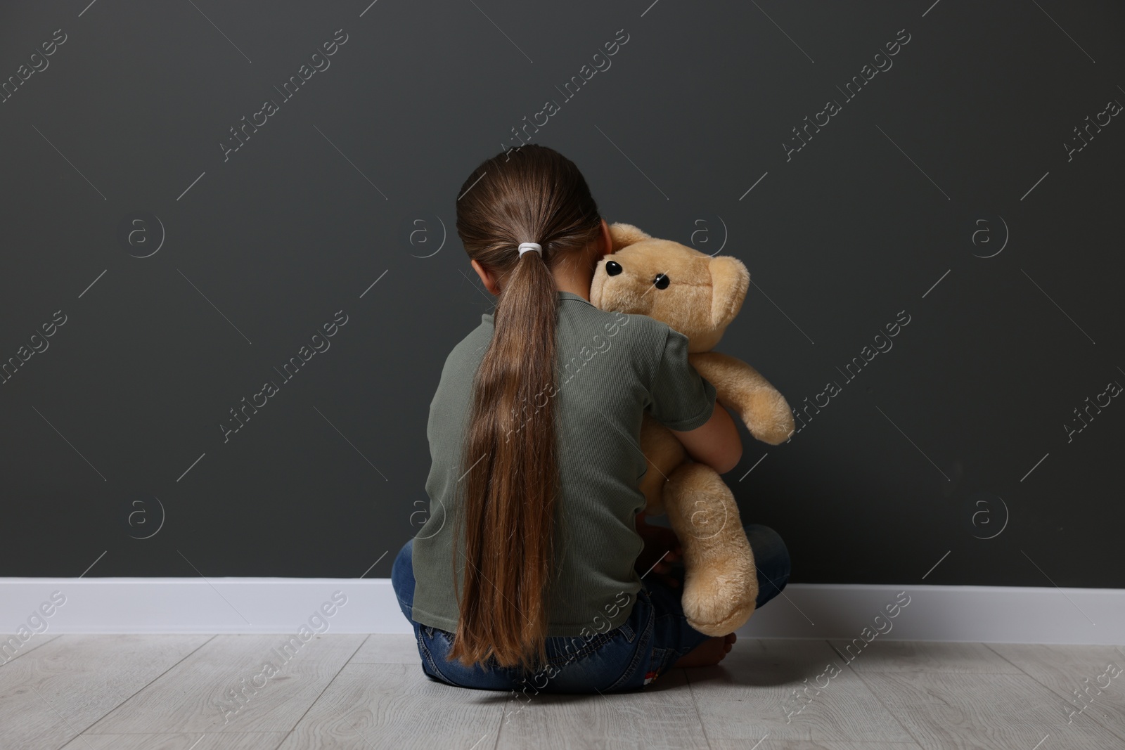 Photo of Child abuse. Upset girl with toy sitting on floor near grey wall, back view