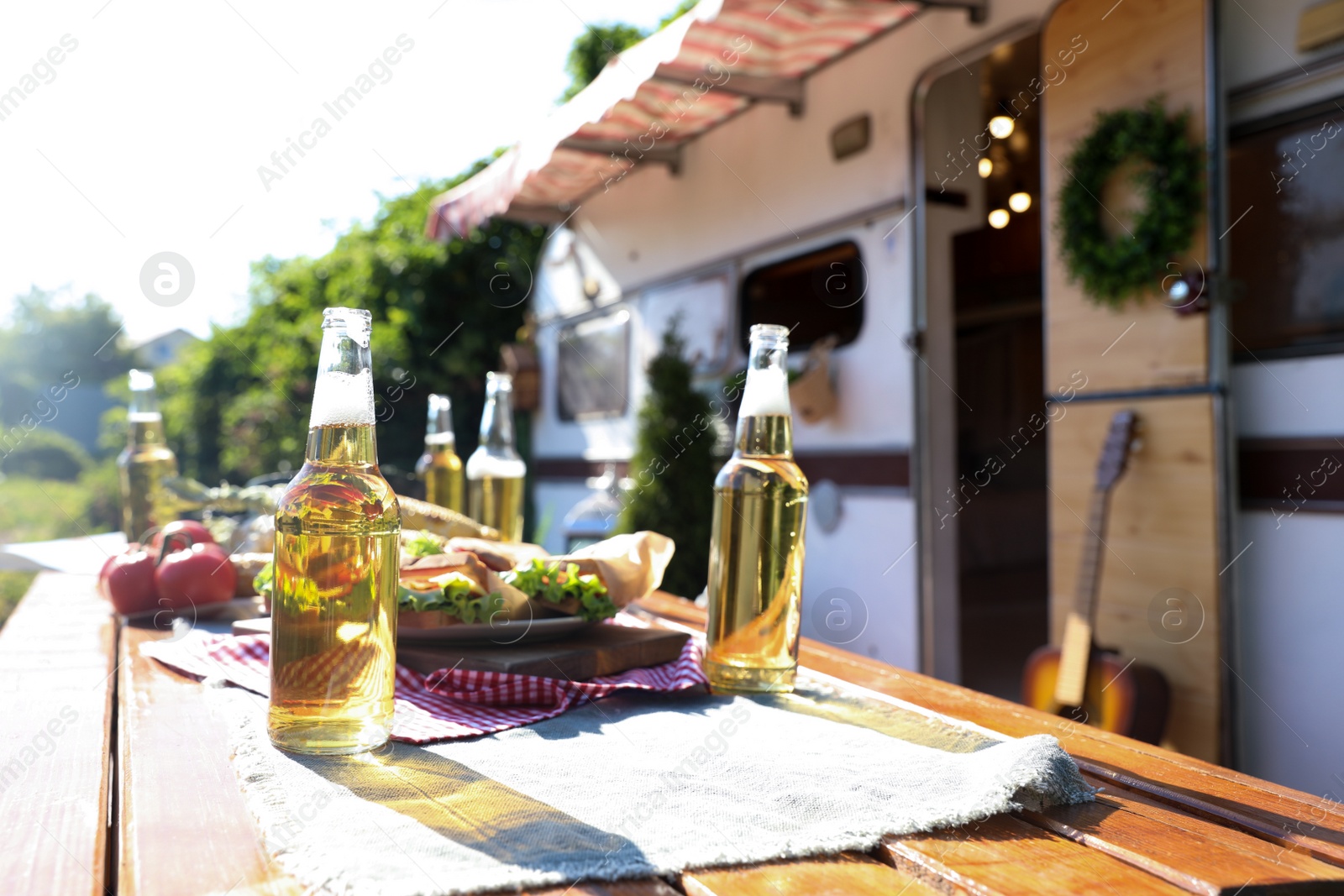 Photo of Delicious sandwiches and bottles of beer on wooden table near motorhome on sunny day. Camping season