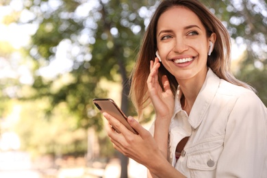 Young woman with wireless headphones and mobile device listening to music in park. Space for text