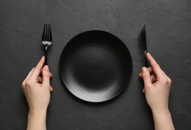 Woman with empty plate and cutlery at black table, top view
