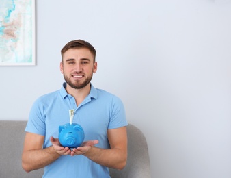 Photo of Young man with piggy bank on sofa in living room. Space for text