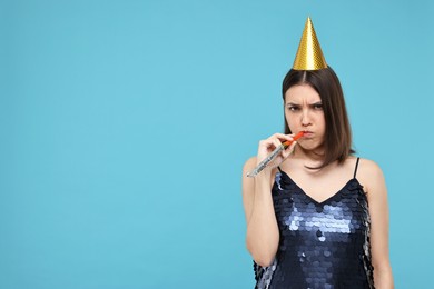 Sad young woman in party hat with blower on light blue background