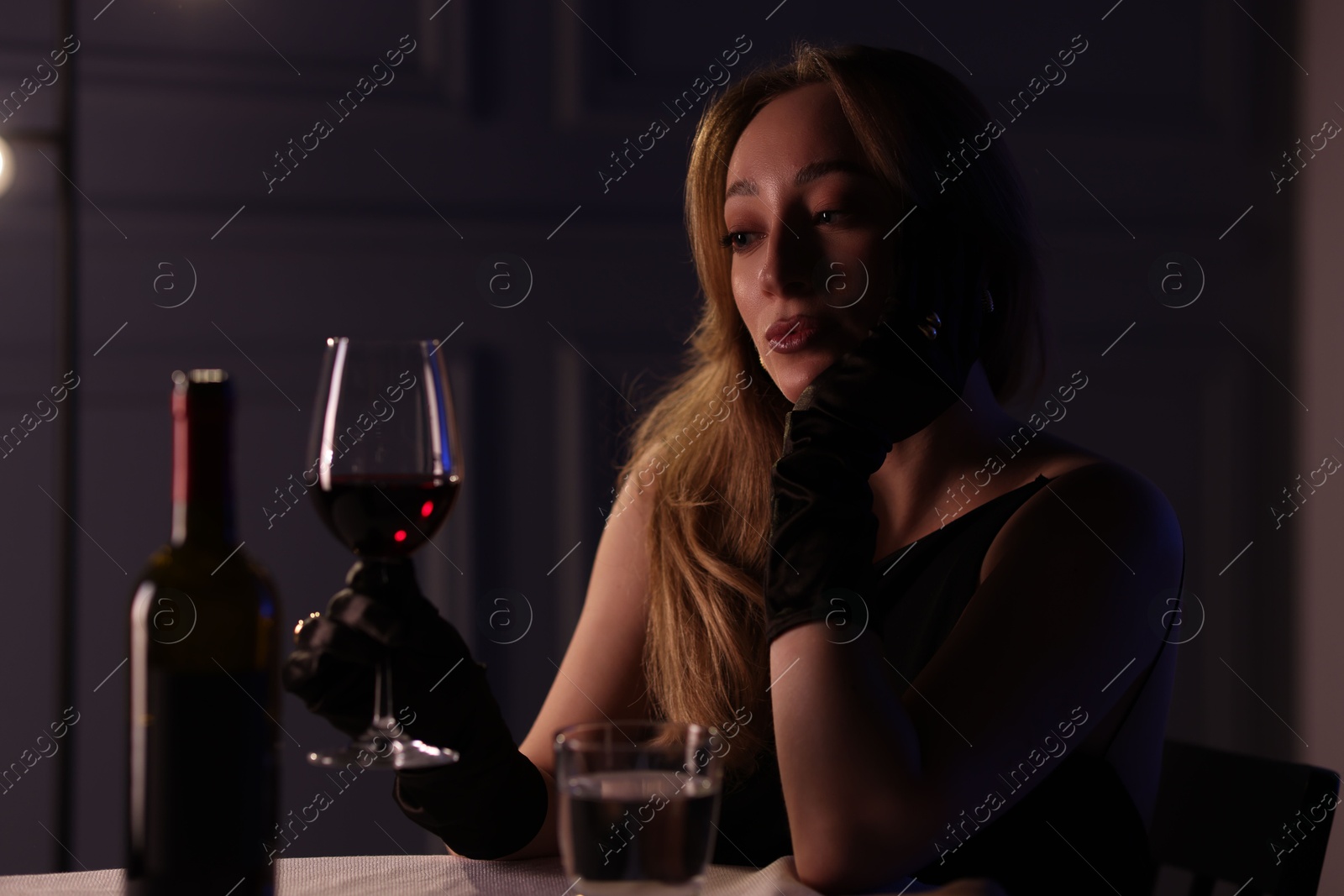 Photo of Elegant young woman with glass of wine at table indoors in evening