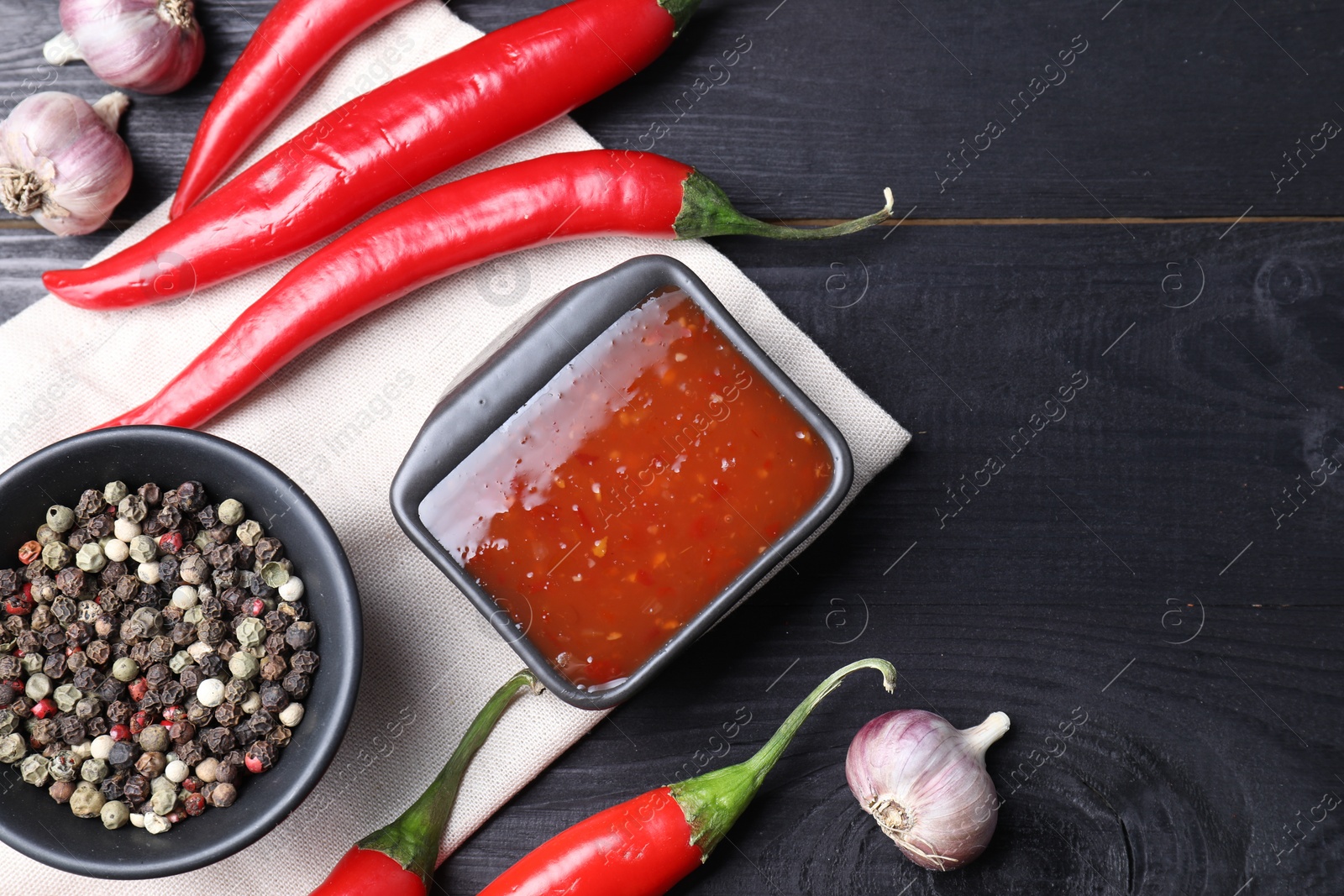 Photo of Spicy chili sauce, garlic, peppers and peppercorns on black wooden table, top view. Space for text