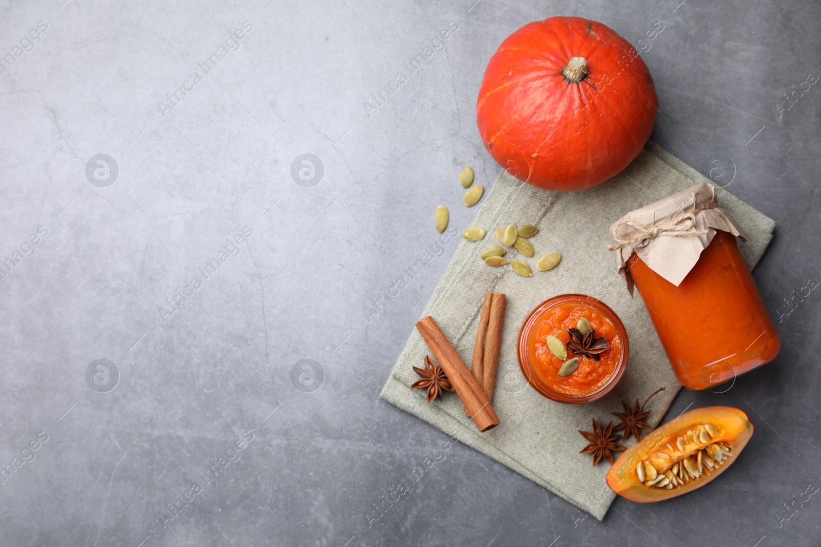 Photo of Jars of pumpkin jam, star anise, cinnamon and fresh pumpkins on grey table, flat lay. Space for text