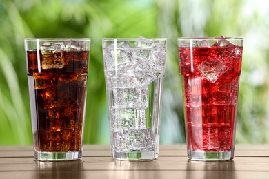 Photo of Glasses of different refreshing soda water with ice cubes on wooden table outdoors