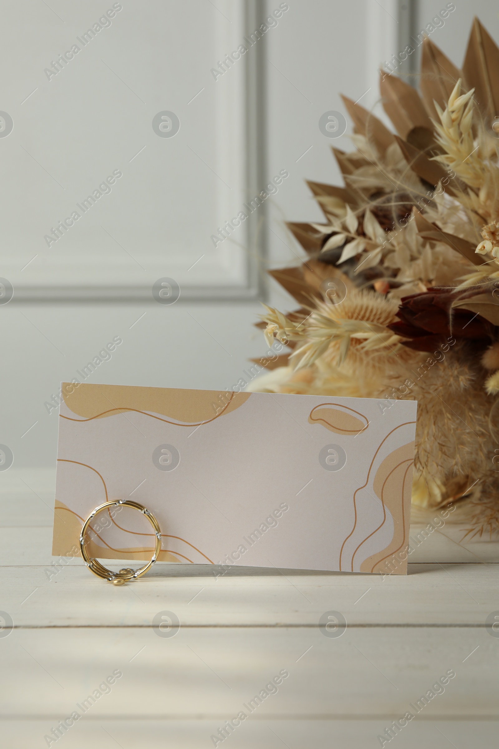 Photo of Blank invitation card, ring, dry leaves and flowers on white wooden table. Space for text