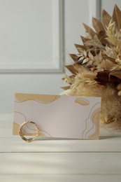 Blank invitation card, ring, dry leaves and flowers on white wooden table. Space for text