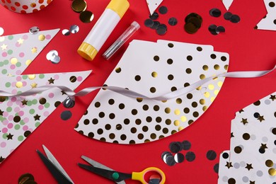 Handmade party hat templates, confetti and tools on red background, flat lay