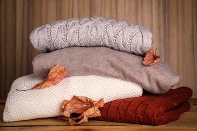 Stack of folded knitted sweaters and autumn leaves on table against wooden background