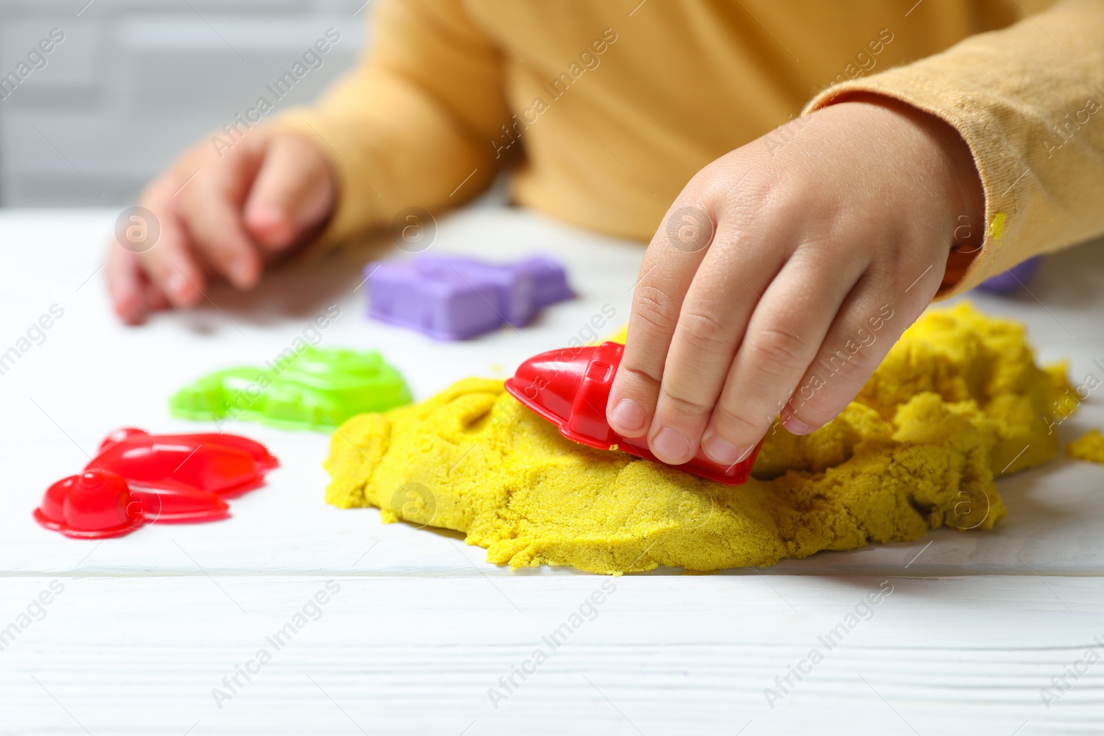 Photo of Little child playing with yellow kinetic sand at white wooden table, closeup