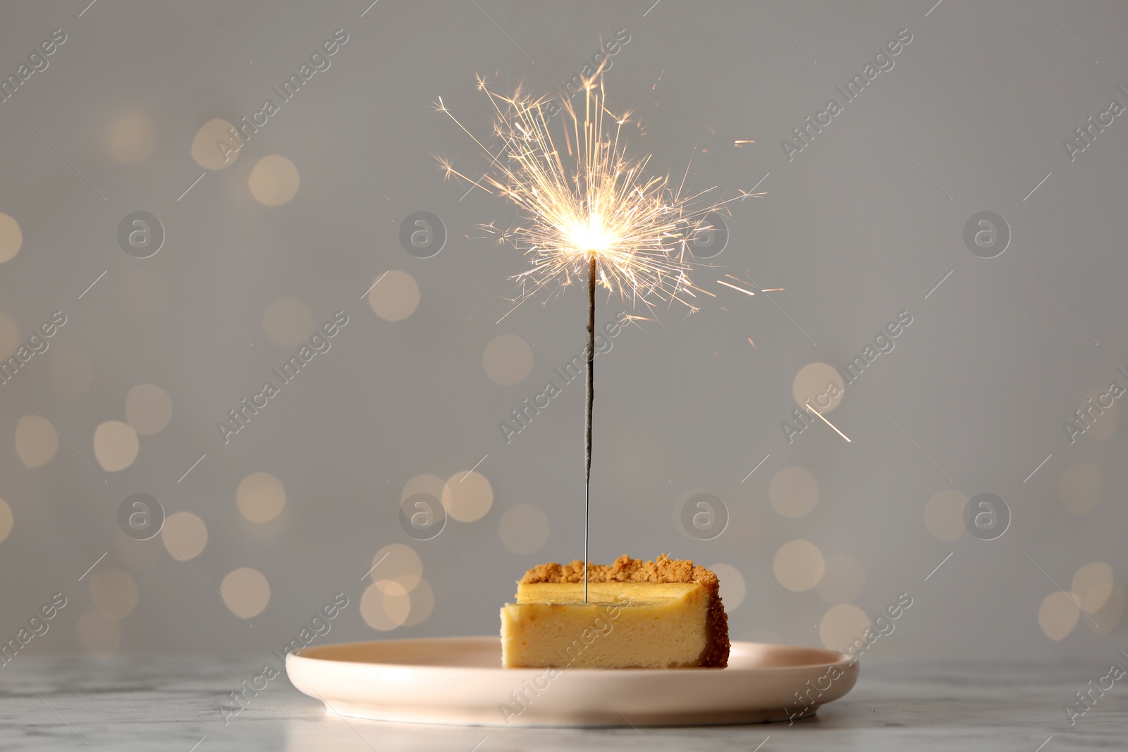 Photo of Piece of delicious cake with burning sparkler on white marble table against blurred lights