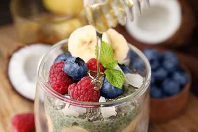 Photo of Pouring honey from dipper onto tasty chia matcha pudding with fruits at wooden table, closeup. Healthy breakfast