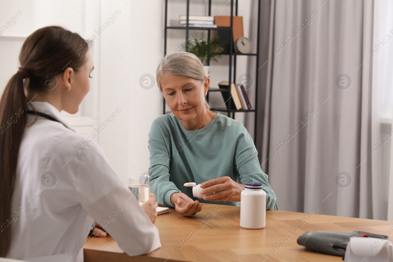 Photo of Young healthcare worker giving glass of water to senior woman with pills at wooden table indoors