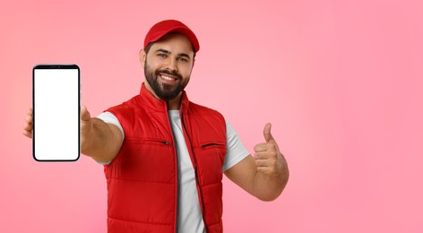 Image of Happy courier holding smartphone with empty screen and showing thumbs up on pink background. Banner design with space for text