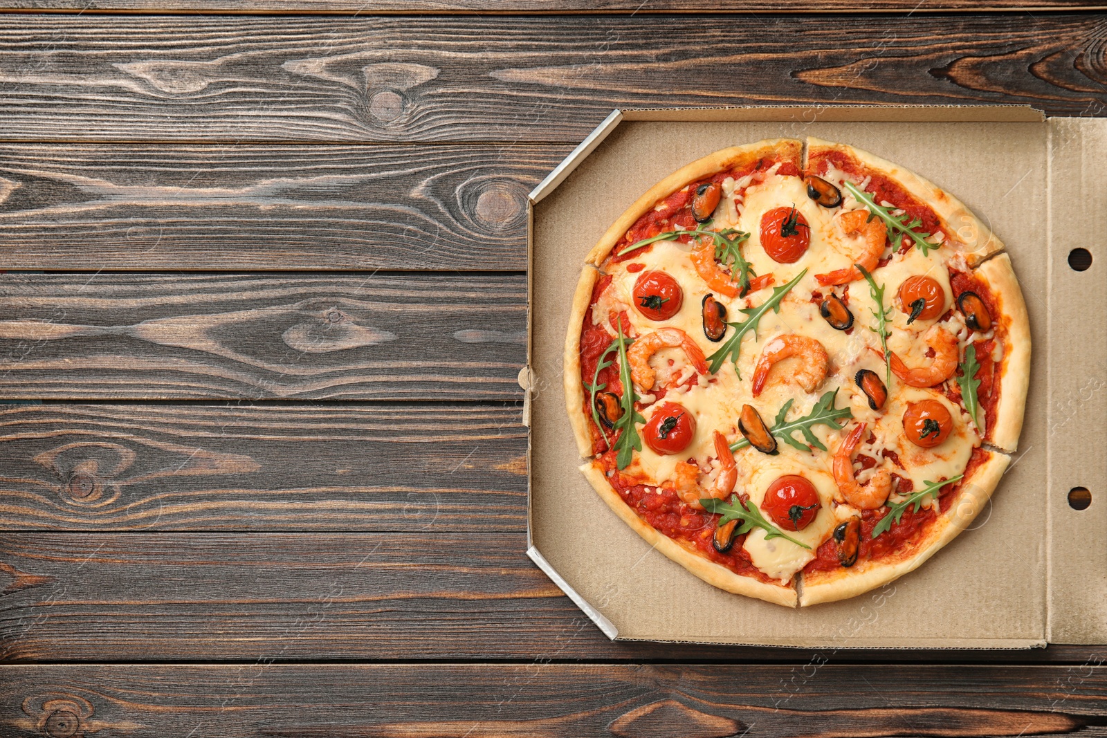 Photo of Delicious seafood pizza in cardboard box on wooden table, top view. Space for text