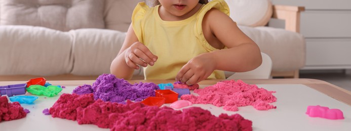 Image of Cute little girl playing with bright kinetic sand at table in room, closeup. Banner design