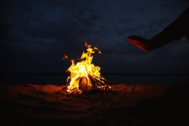 Woman warming hands near burning firewood on beach at night, closeup