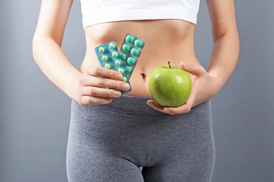 Slim woman holding weight loss pills in blister packs and apple against gray background, closeup