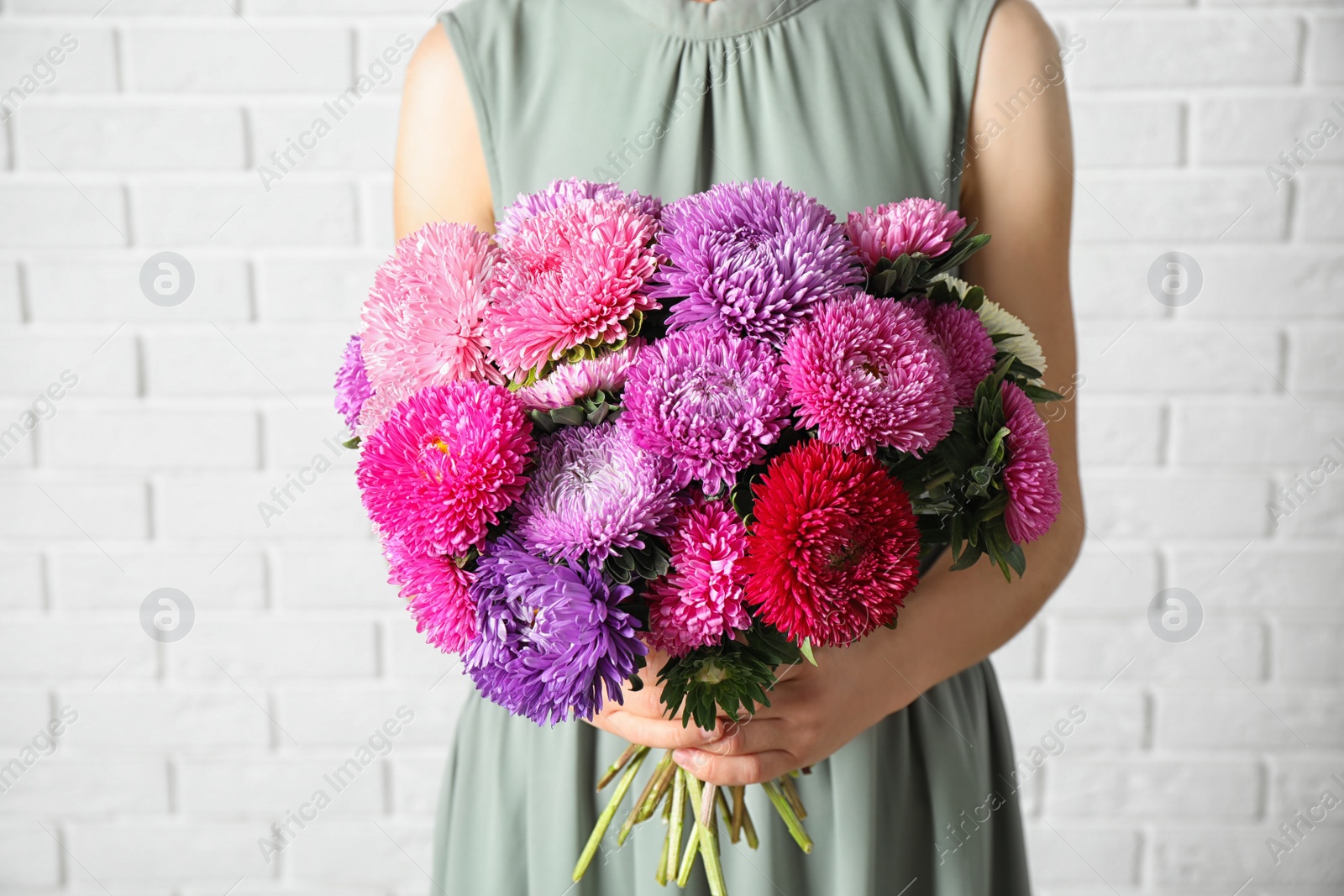 Photo of Woman holding bouquet of beautiful aster flowers against white brick wall, closeup