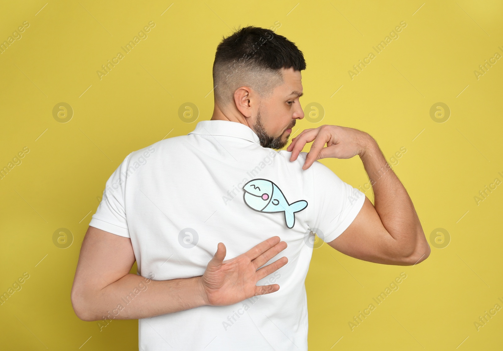 Photo of Man with paper fish on back against yellow background. April fool's day