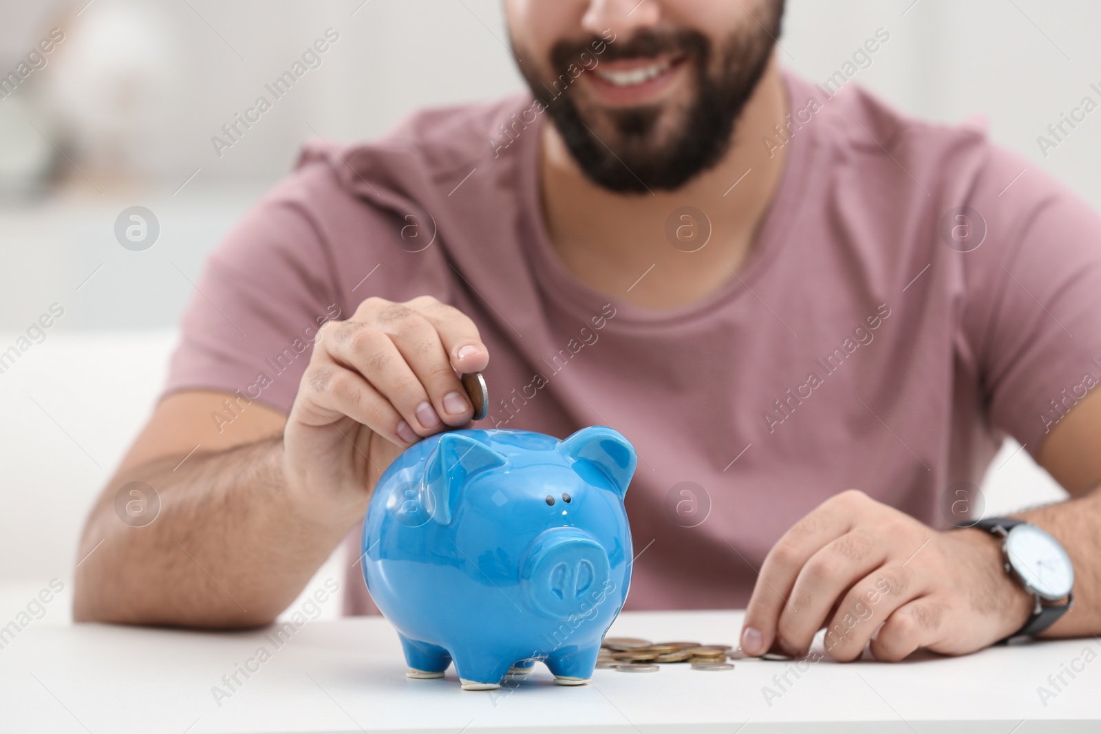 Photo of Happy young man putting money into piggy bank at white table indoors, closeup
