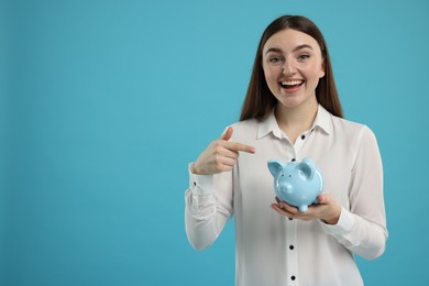 Excited woman pointing at piggy bank on light blue background, space for text