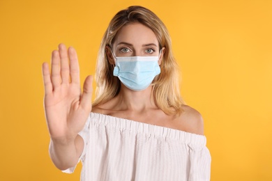 Photo of Woman in protective face mask showing stop gesture on yellow background. Prevent spreading of coronavirus