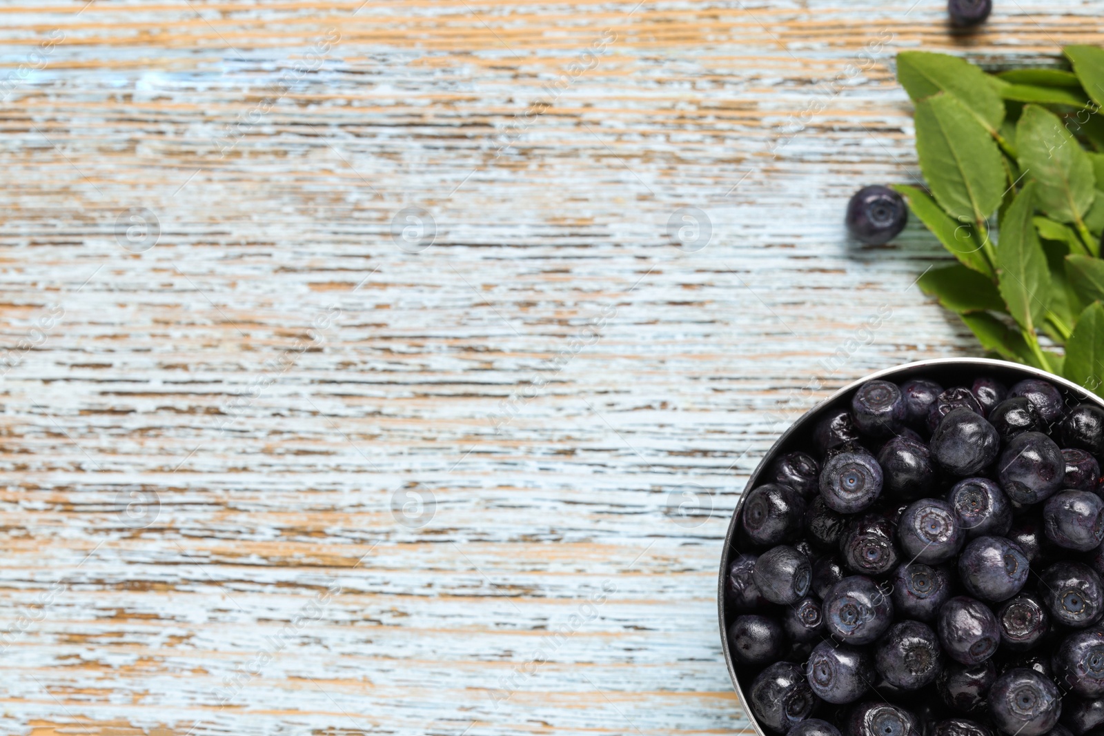 Photo of Ripe bilberries in bowl and sprigs with leaves on wooden rustic table, flat lay. Space for text