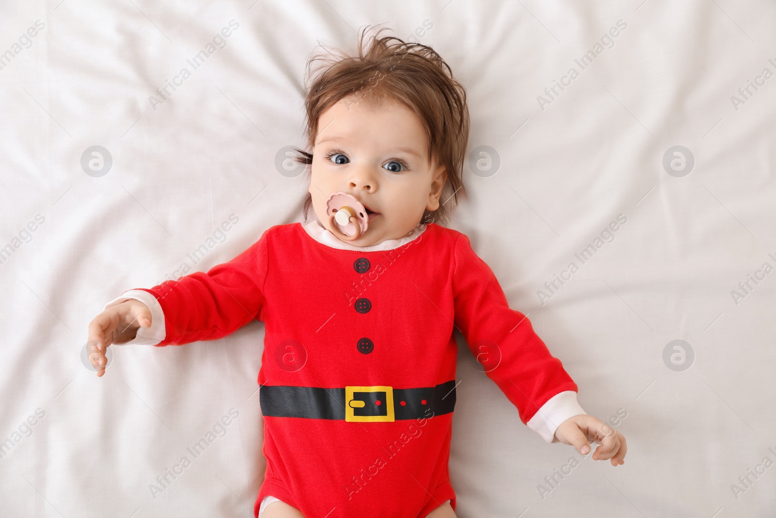 Photo of Cute baby wearing festive Christmas costume with pacifier on white bedsheet, top view