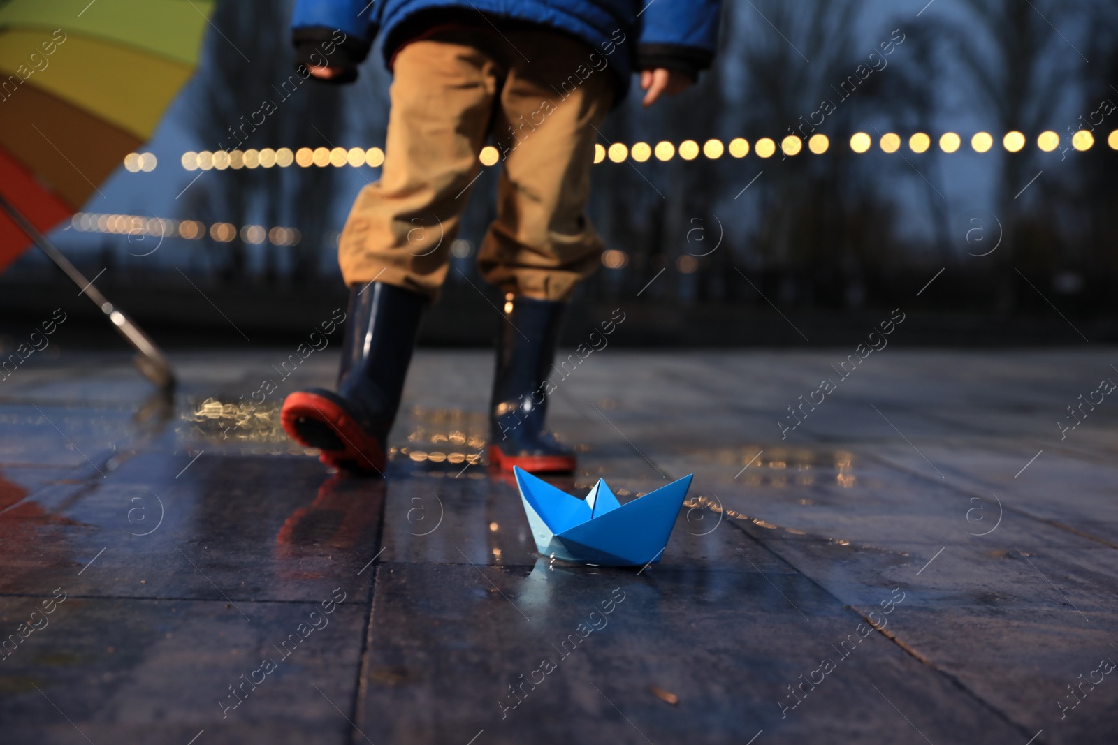 Photo of Little boy outdoors, focus on paper boat in puddle