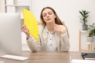 Photo of Businesswoman waving yellow hand fan to cool herself at table in office