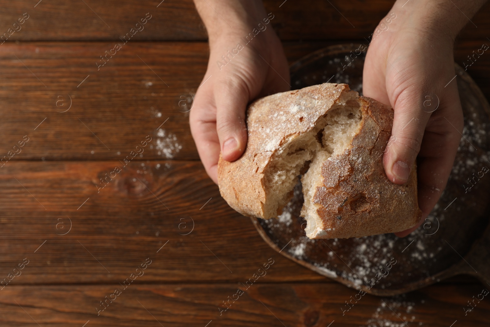 Photo of Man breaking loaf of fresh bread at wooden table, top view. Space for text