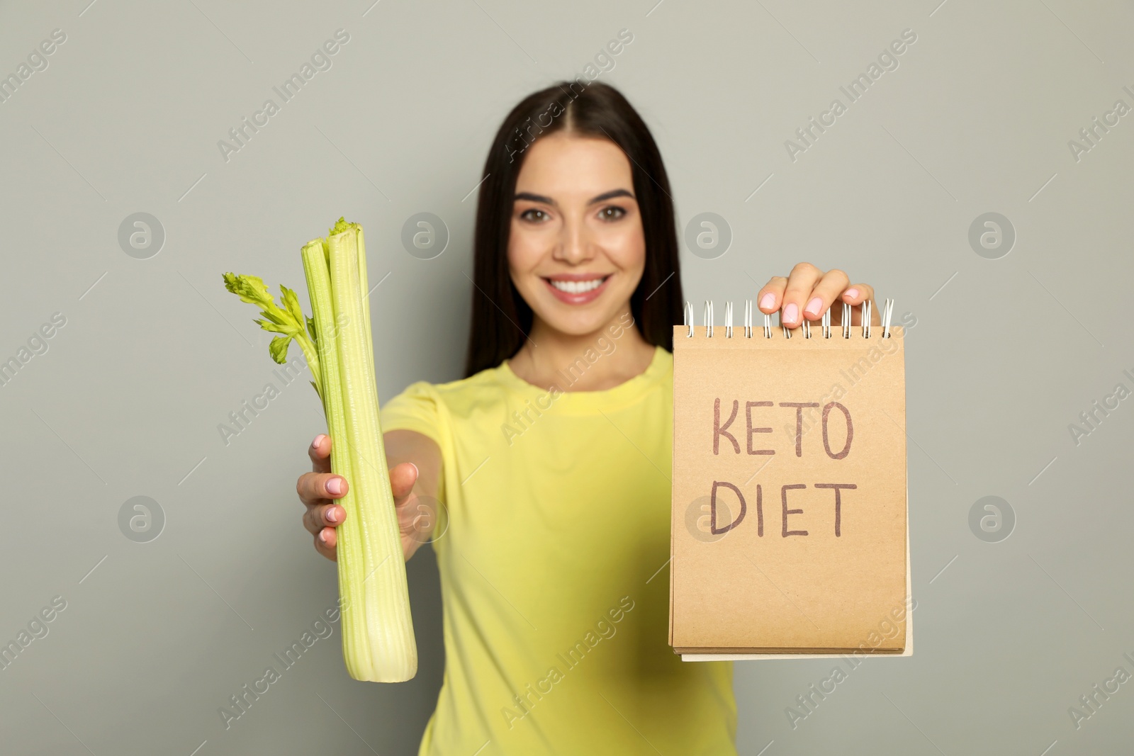 Photo of Happy woman holding celery and notebook with words Keto Diet against light grey background, focus on hands