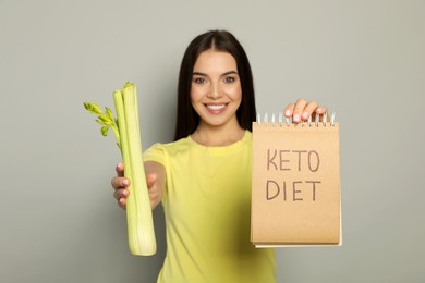 Happy woman holding celery and notebook with words Keto Diet against light grey background, focus on hands