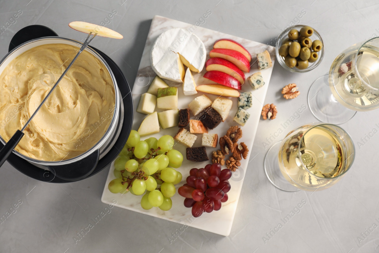 Photo of Fork with piece of apple, melted cheese in fondue pot, wine and snacks on grey table, flat lay