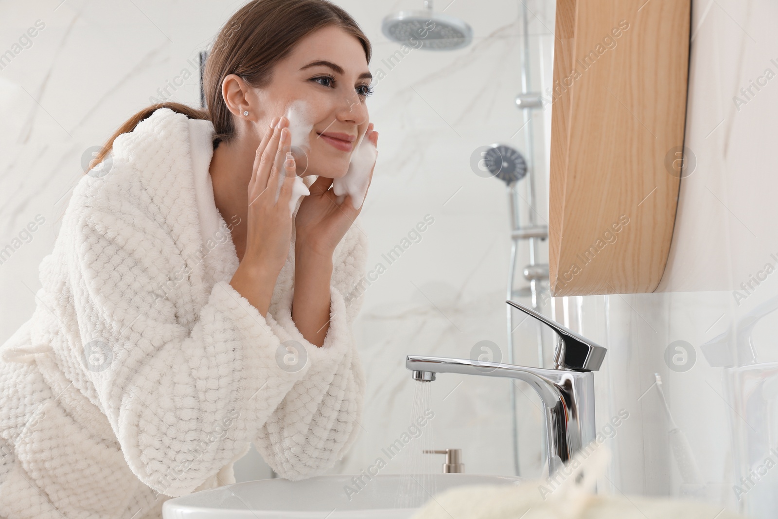 Photo of Young woman applying cleansing foam onto her face in bathroom