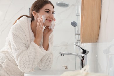 Young woman applying cleansing foam onto her face in bathroom
