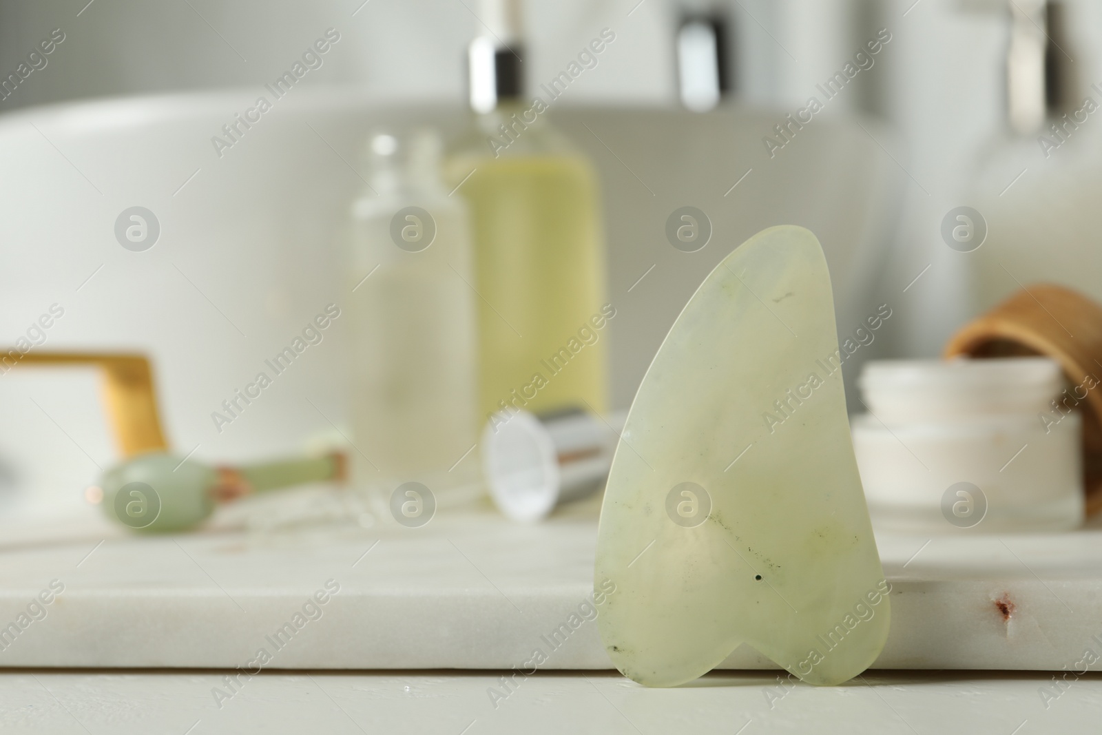 Photo of Jade gua sha tool and toiletries on white countertop in bathroom, closeup. Space for text