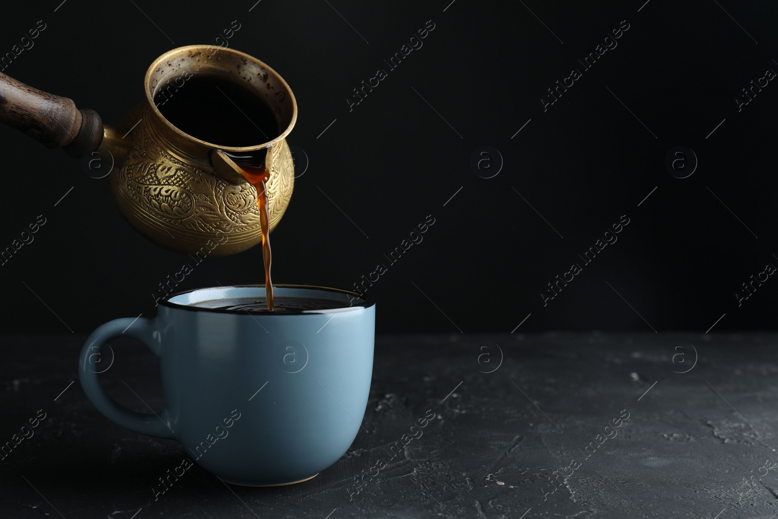 Photo of Turkish coffee. Pouring brewed beverage from cezve into cup at black table. Space for text