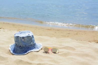 Photo of Jeans hat and sunglasses on sand near sea, space for text. Beach accessories