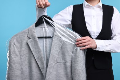 Dry-cleaning service. Woman holding jacket in plastic bag on light blue background, closeup