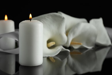 Photo of Burning candle and white calla lily flowers on black mirror surface in darkness, closeup with space for text. Funeral symbols