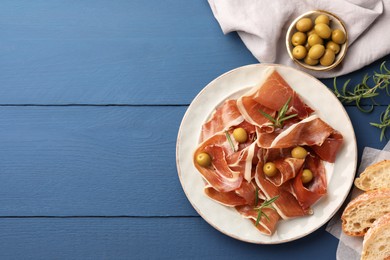 Photo of Slices of tasty cured ham, rosemary, bread and olives on blue wooden table, flat lay. Space for text