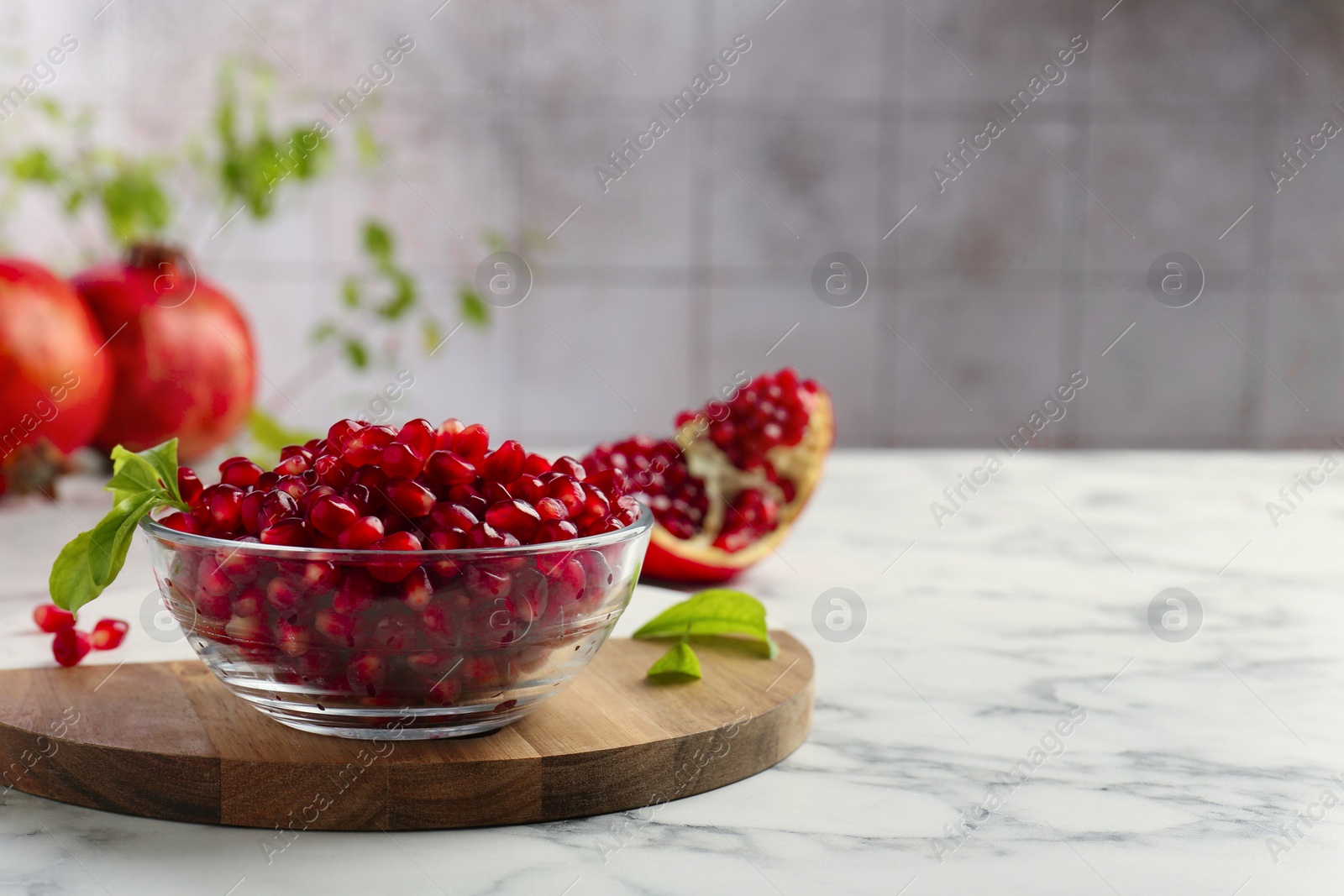 Photo of Ripe juicy pomegranate grains in bowl and green leaves on white marble table, space for text