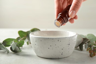 Photo of Woman dripping eucalyptus essential oil from bottle into bowl at white table, closeup