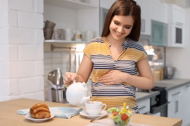 Photo of Young pregnant woman pouring tea into cup at table in kitchen