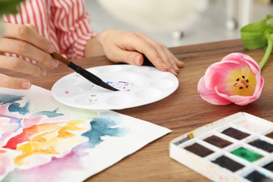 Photo of Woman painting flowers with watercolor at wooden table, closeup. Creative artwork