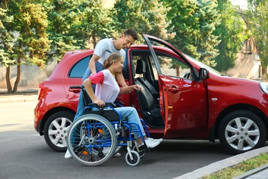 Young man helping disabled woman in wheelchair to get into car outdoors