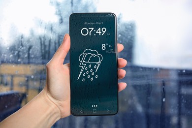 Image of Woman checking weather using app on smartphone near wet window, closeup. Data and illustration of cloud with lightning and rain on screen